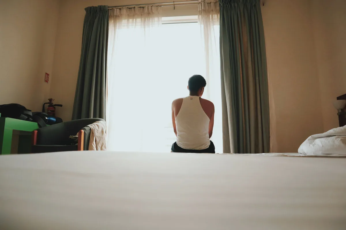 Person seated on a bed facing a bright window in a quiet room